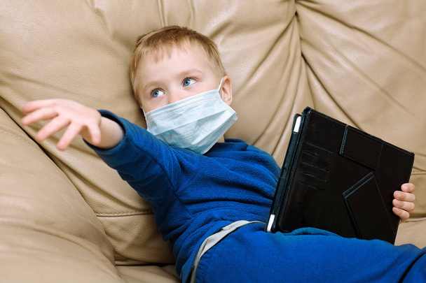 Cute baby boy with bright blue eyes in medical sanitary mask, with pad digital tablet. Wearing blue pajamas, emotional face expressiion. Illness or virus concept, indoors, copy space. - Foto, Imagem