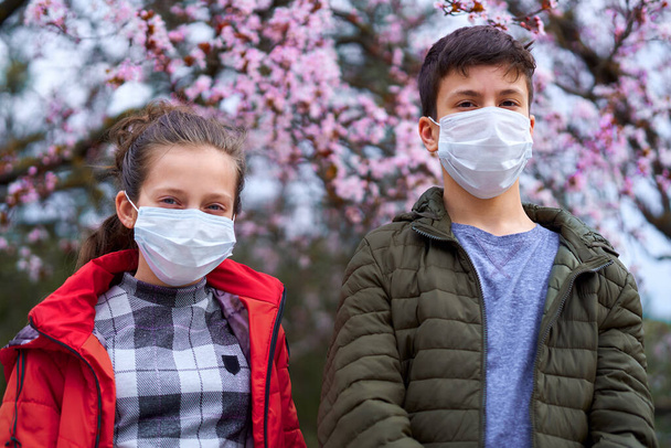 Mädchen und Jungen mit Gesichtsmaske sind in der Stadt draußen, blühende Bäume, Frühlingszeit, Blütezeit - Allergiekonzept und Gesundheitsschutz vor staubiger Luft - Foto, Bild