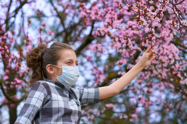 девушка в маске для лица находится в городе под открытым небом, цветущие деревья, весенний сезон, время цветения - концепция аллергии и защиты здоровья от пыльного воздуха
 - Фото, изображение