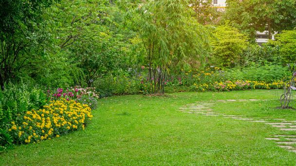 Césped de hierba verde en un jardín con patrón aleatorio de piedra escalonada de hormigón gris, planta con flores, arbustos, árboles en el patio trasero bajo el sol de la mañana con buen cuidado paisajismo en un parque público
   - Foto, imagen