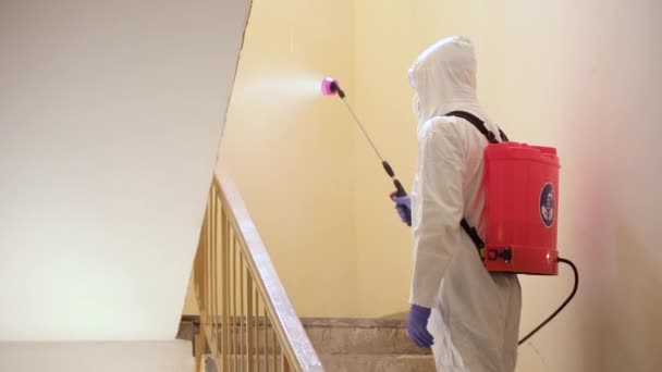 Een man in beschermende apparatuur desinfecteert met een spuitbus in het gebouw. Oppervlaktebehandeling als gevolg van coronavirus covid-19 ziekte. Een man in een wit pak desinfecteert het huis met een spuitpistool. Viruspandemie - Video