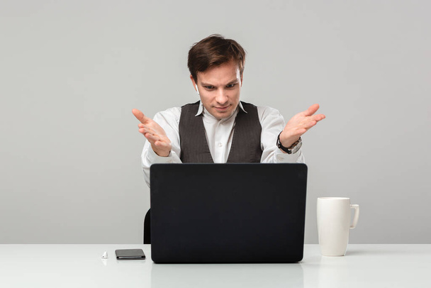 Zakenman in een wit t-shirt en grijs vest is geschokt van het controleren van informatie op een laptop aan de witte tafel. Dagelijkse zakelijke routine. - Foto, afbeelding