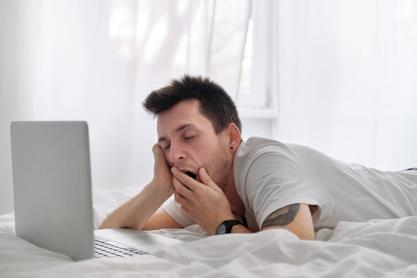Der gutaussehende junge Mann im weißen Hemd und Pyjama auf einem Bett mit weißer Bettwäsche blickt aufgebracht auf den Bildschirm eines Laptops. Morgen eines freien Mitarbeiters - Foto, Bild