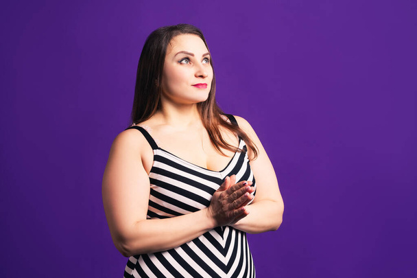 Pensive plus size model in striped dress, fat woman on purple background, body positive concept - Фото, изображение