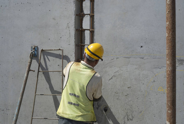 Ein Bauarbeiter installiert eine Betonfertigteilwandkonstruktion mit Stahlstange auf der obersten Etage des Gebäudes, trägt orangefarbenen Schutzhelm und grüne Weste, den Stützstahl und eine feste Leiter links - Foto, Bild