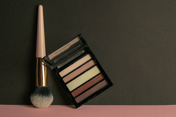 Un pincel de maquillaje y un soporte de sombra meicap sobre una mesa rosa apoyada contra una pared negra
. - Foto, Imagen