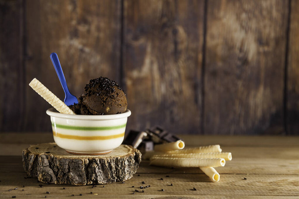 Dwie kulki lodów czekoladowych na ceramicznej misce z niebieską plastikową łyżeczką i herbatnikiem na kawałku drewna i różne herbatniki na drewnianym tle. Przestrzeń kopiowania - Zdjęcie, obraz