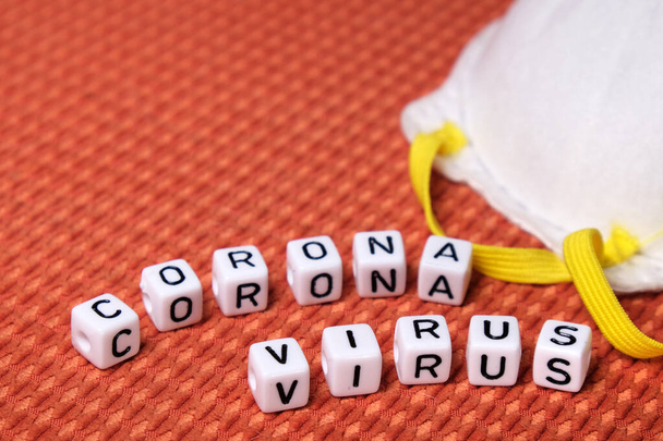 Weiße Schutzmaske und Coronavirus-Wort aus weißen Blöcken auf einem Texturhintergrund. Kopierraum, Ansicht von oben. - Foto, Bild