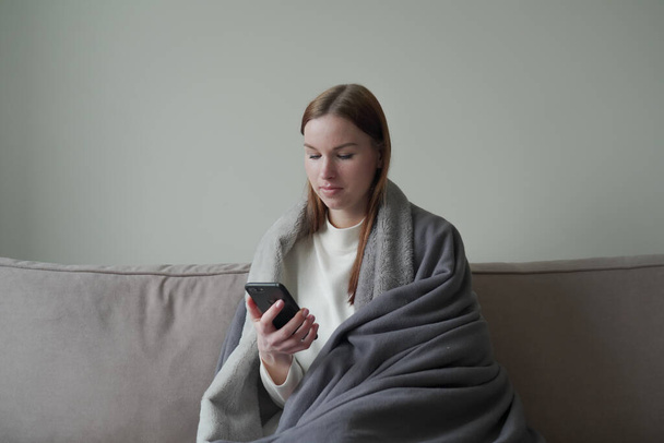 Beautiful young girl sitting on a sofa in a medical mask and looking at the phone. It works on quarantine. Orvi freelancer, coronovirus, flu, acute respiratory infections. - Foto, Imagem