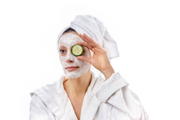 Young beautiful girl with a white cosmetic mask on her face holds a cucumber in her hand. - Foto, Imagem