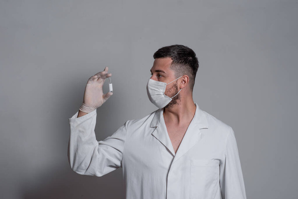 Mies valkoisessa lääketieteellisessä puvussa, naamio kasvoillaan ja leikkauskäsineet katsoo lasipurkkia rokotetta valkoisella tai harmaalla taustalla.
 - Valokuva, kuva