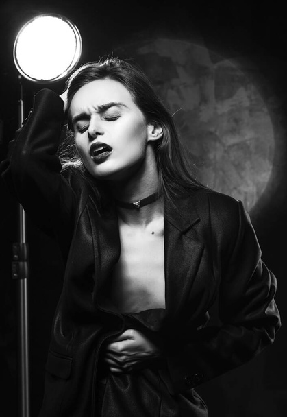 Beautiful slim braless girl, wearing a unbuttoned black blazer, sensually frowns and touches her hair with her hand on a dark background, posing next to a light lamp. Advertising monochrome design - Photo, image