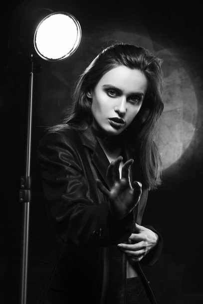 Beautiful slim girl model with red lips, wearing a black blazer, fearfully protects herself with her hand on a dark background, next to the light lamp. Advertising, trendy, monochrome design - Фото, изображение