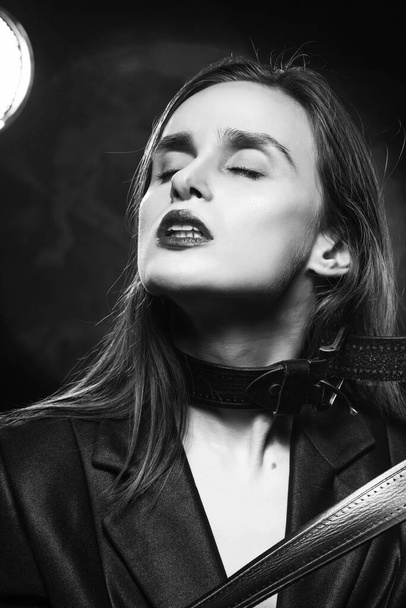 Braless girl with red lips, wearing a unbuttoned blazer and a leather belt tightened around the neck, which she pulls with her hands,  on the dark background, next to a light lamp. Monochrome design - Fotografie, Obrázek