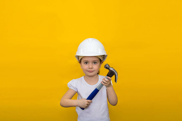 happy little girl in a construction white helmet holding a blue hammer on a yellow background. concept of renovation. - Foto, afbeelding