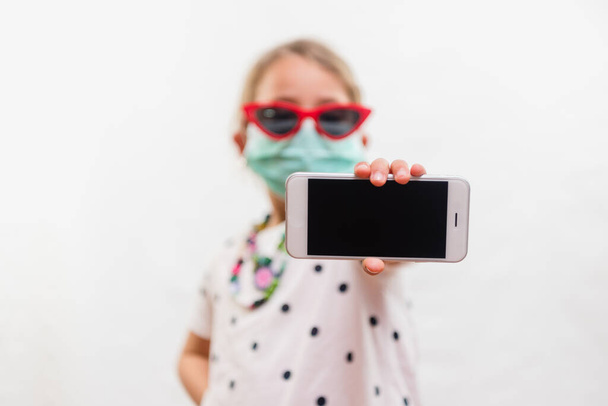 Pieni vaalea tyttö yllään kertakäyttöinen naamio ja aurinkolasit tilalla matkapuhelin valkoisella taustalla
 - Valokuva, kuva