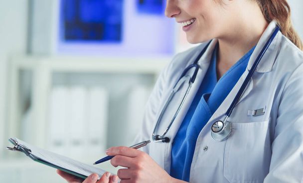 Doctor with a stethoscope, holding a notebook in his hand. Close-up of a female doctor filling up medical form at clipboard while standing straight in hospital - 写真・画像