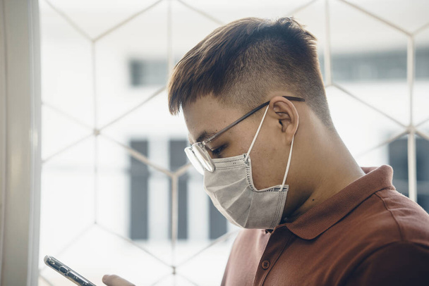 Close up of one young Asian man wearing a white surgical face mask and using a mobile phone during new type Coronavirus Covid-19 pneumonia outbreak and pm 2.5 smog air pollution crisis in big city - Фото, зображення