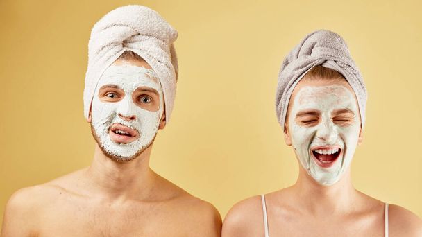 Young friends guy and girl with funny expressive emotions, towel on head and face mask isolated on a yellow background, skin care concept - Foto, afbeelding