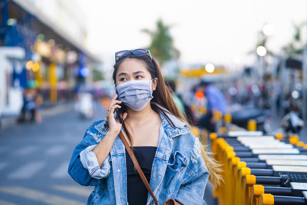 young woman wearing surgical mask on street while using mobile phone. Portrait of young woman wearing a protective mask to prevent germs, toxic fumes, and dust. Prevention of bacterial infection Corona virus or Covid 19. - Фото, изображение