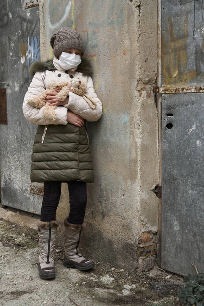 koncept epidemie a karantény - dívka s obličejovou maskou a roztomilou hračkou sama na ulici ve městě - Fotografie, Obrázek