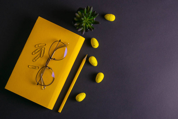 Sárga ceruza, sárga notebook szemüveg, csapok, klip papír és sárga húsvéti tojás fekete alapon. - Fotó, kép