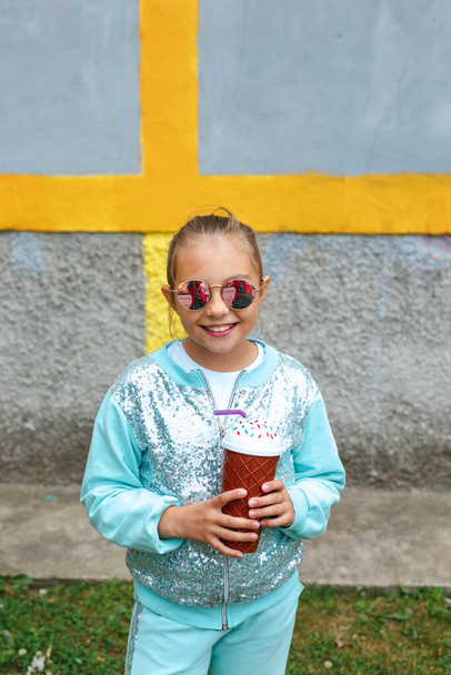 Little stylish girl in sunglasses is drinking milk cocktail over city background - Foto, Bild