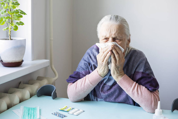 Ηλικιωμένη λευκή γυναίκα με συμπτώματα γρίπης, στο Home Quarantine. Στο τραπέζι είναι η προστατευτική μάσκα, χάπια. Concept - Επικίνδυνος ιός για Ηλικιωμένους, Αυτοαπομόνωση. - Φωτογραφία, εικόνα
