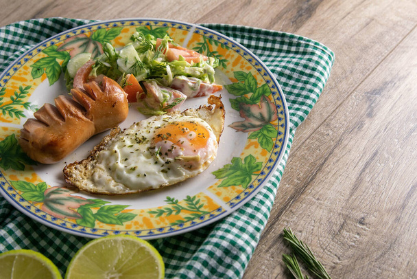 Anglická snídaně, míchaná vejce s klobásou a zelným salátem, vedle je limetka rozříznutá vejpůl, talíř stojí na dřevěném stole. - Fotografie, Obrázek