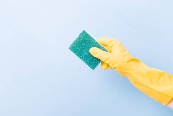 a hand in a yellow rubber glove holds a yellow sponge with a green rough side for cleaning complex dirt, blue background copy space, cleaning concept, home care, spring cleaning - Фото, изображение