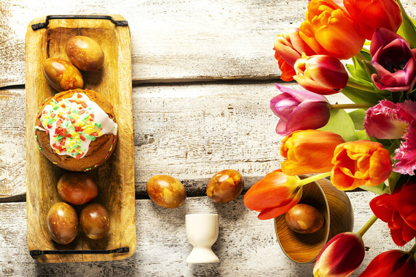 Exposition of Easter eggs in wooden plate with beautiful tulip flowers and Easter cake on white wooden background. - Photo, image