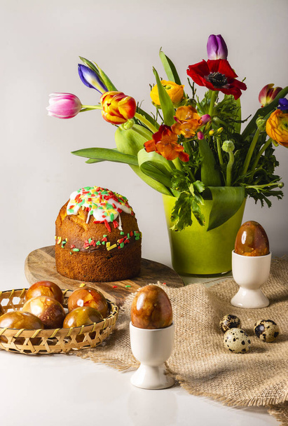 Ahşap tabakta Paskalya yumurtaları, güzel lale çiçekleri ve beyaz ahşap arka planda Paskalya pastası sergisi.. - Fotoğraf, Görsel