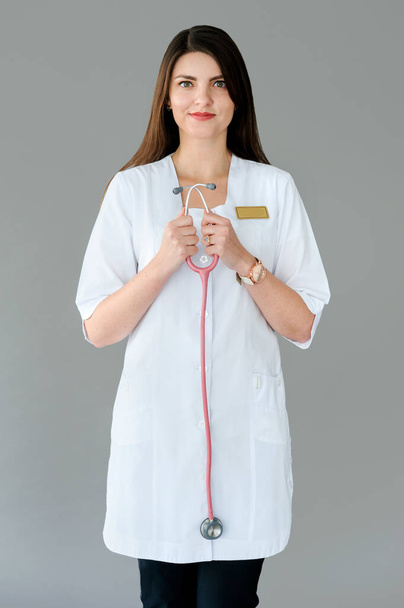 beautiful young female doctor with phonendoscope stands on gray background - Фото, зображення