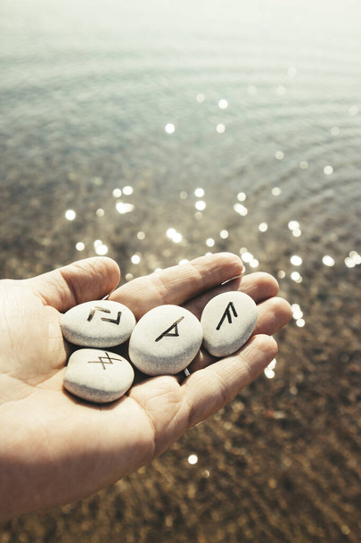 Hand mit Runensteinen mit schwarzen Symbolen für den Blick in die Zukunft, schönes sonniges Bokeh-Meerwasser im Freien auf dem Hintergrund. - Foto, Bild