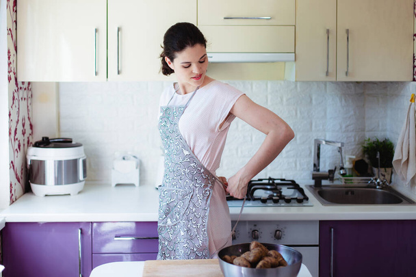 Young woman in kitchen knot an apron on her back - Фото, изображение
