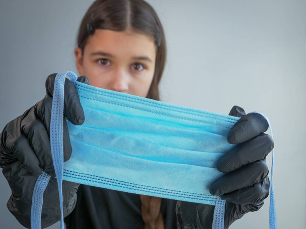 Siyah lateks eldivenli genç bir bayan kameraya bakıyor ve ön planda mavi koruyucu bir tıbbi maske tutuyor. Koronavirüs pandemisi sırasında antiviral koruma. - Fotoğraf, Görsel