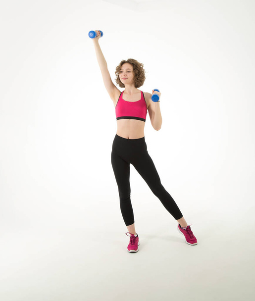 Full length, young athletic woman with dumbbells in her hands doing fitness exercises, standing on a white background. - Фото, изображение
