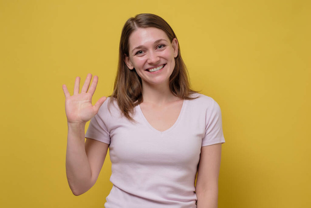 Attractive friendly looking young woman smiling happily, saying Hello, Hi or Bye, waving hand. Showing five fingers. Studio shot on yellow wall - Photo, Image