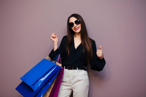 Portré izgatott csinos lány visel blúz, fehér nadrág és napszemüveg gazdaság bevásárlótáskák elszigetelt kék háttér. - Fotó, kép