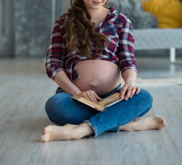 Schwangere sitzt mit einem Buch auf dem Boden. Konzept der glücklichen Mutterschaft - Foto, Bild