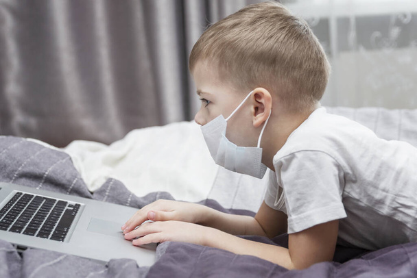 A boy in a medical mask sits in front of a laptop at home in bed. Distance learning. Quarantine in the context of a coronavirus pandemic. - Φωτογραφία, εικόνα