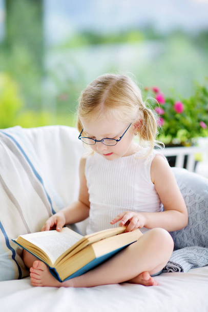 Little girl reading a book in living room. Smart schoolgirl doing her homework after school. Education and distance learning for kids. Homeschooling during quarantine. Stay at home entertainment. - Zdjęcie, obraz