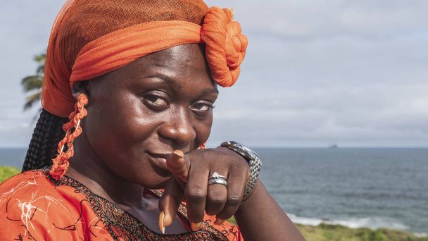 Mulher africana com vestido laranja e capacete de Gana olhando para a câmera e apontando o dedo para o fotógrafo
 - Foto, Imagem