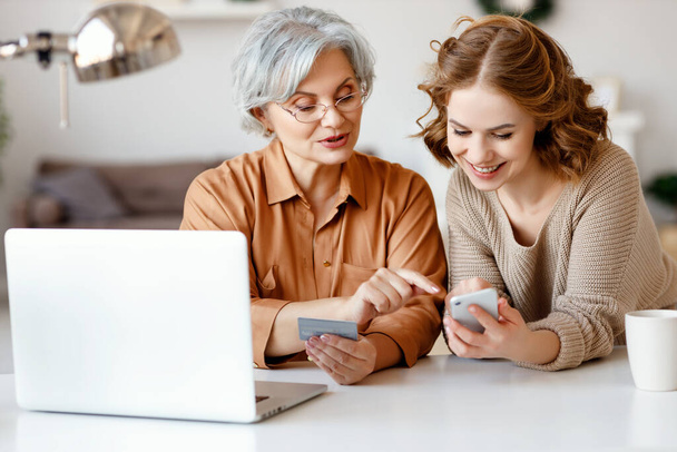Young granddaughter smiling and showing smartphone to elderly lady with credit card while shopping online at home togethe - Фото, изображение