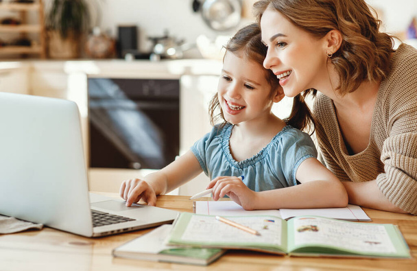 Positive young woman helping daughter in searching information for homework on internet while sitting together at table with laptop at hom - Foto, Imagen