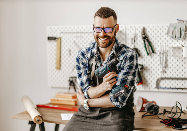 Pozitív fiatal szakállas férfi kötényben és szemüvegben a kamerába néz és mosolyog miközben fúróval a kezében áll a modern worksho-ban - Fotó, kép