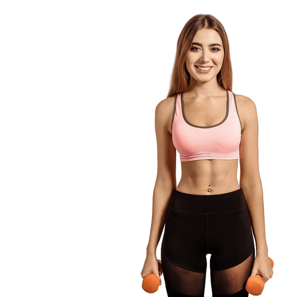 Beautiful, young woman standing in a sportswear, top and leggings. Tight, athletic body, thin, flat stomach. Holding dumbbells in his hands. - Photo, image