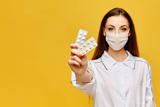 Kobieta-lekarz w mundurze i ochronnej masce medycznej trzymając tabletki w ręce i pozowanie na żółtym tle, przestrzeń kopiowania, izolowane. Koncepcja opieki zdrowotnej - Zdjęcie, obraz
