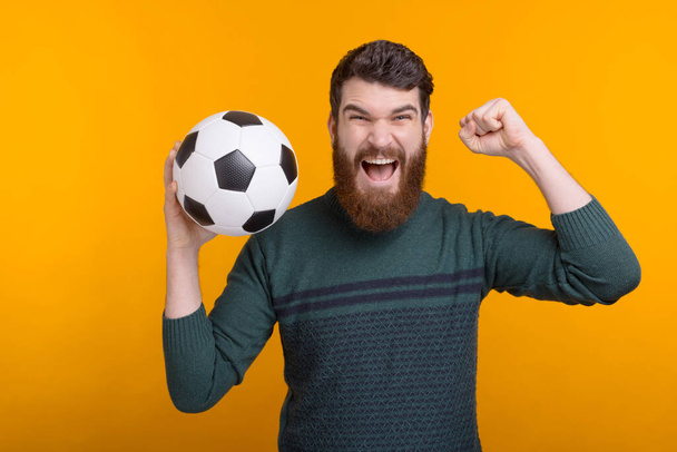 Волнующий бородатый мужчина держит мяч в руке, крича и делая жест победителя на желтом фоне
 - Фото, изображение