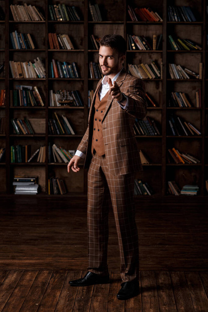 portret van een jonge bebaarde zakenman in een bruin pak tegen de achtergrond van de bibliotheek. - Foto, afbeelding